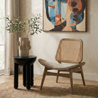 An abstract framed canvas print with geometric shapes in blue, beige, brown, and black, displayed on a textured beige wall in a styled room setting with a wooden chair, a black side table, and a potted plant.