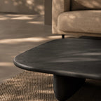 A dark charcoal-colored wooden coffee table with a rounded organic edge and a thick sculptural base, set on a textured beige rug in a modern living room with dappled sunlight.