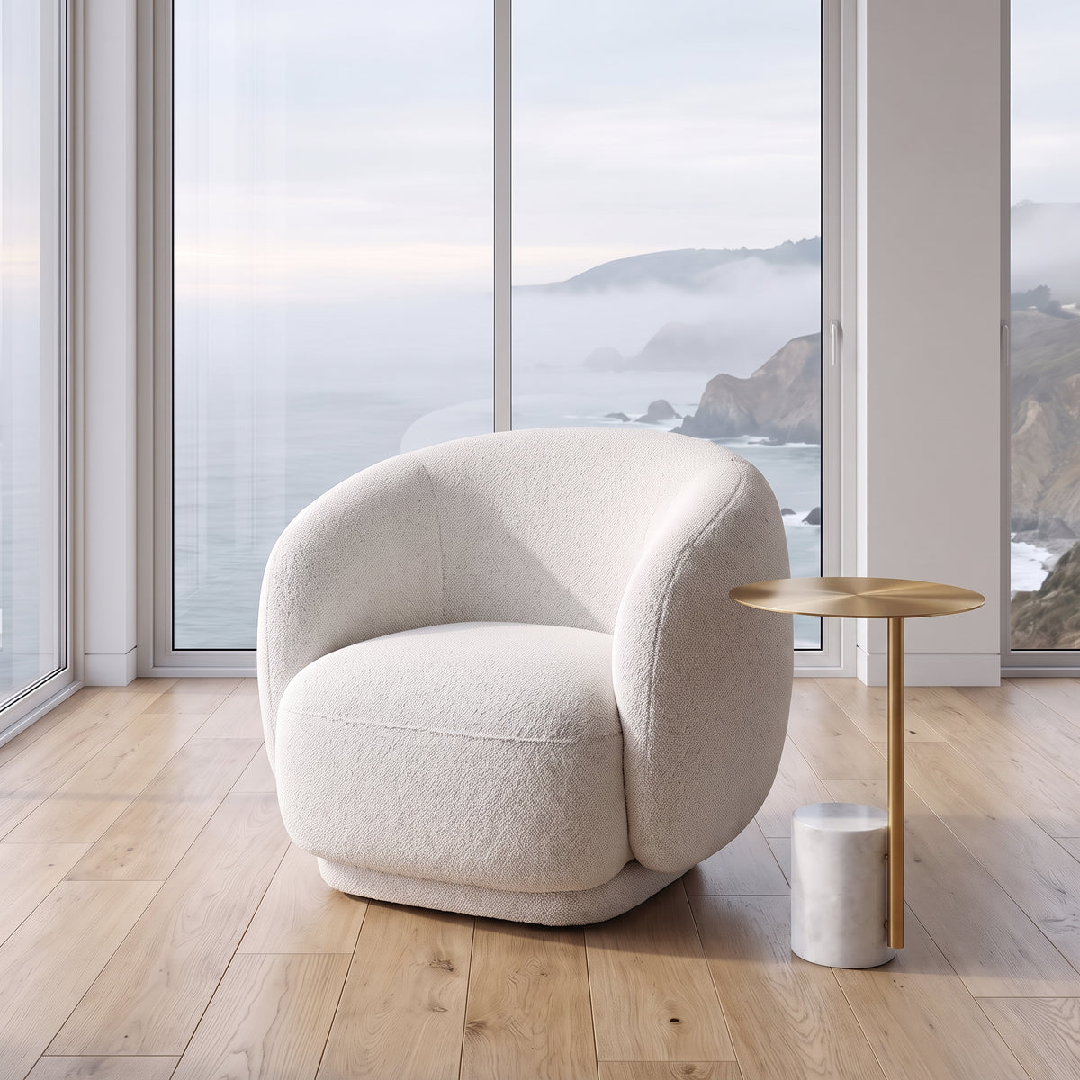 A modern round armchair upholstered in textured cream bouclé fabric, positioned in a bright room with a wooden floor and a small round gold side table next to it. Large windows behind the chair offer a scenic view of a misty coastal ocean.